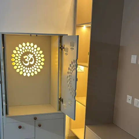 White pooja unit with a jaali-cut door panel and a backlit Om medallion inside, beside open display shelving with cove lighting.