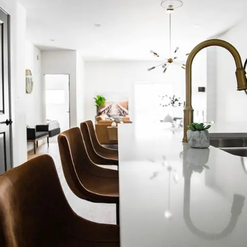 White quartz kitchen island with a brass gooseneck faucet and tan leather counter stools, opening onto a sunlit living room beyond