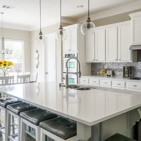 White shaker kitchen with a large quartz island, glass globe pendant lights, and a grey subway-tile backsplash, opening onto a dining nook.