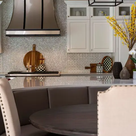 White shaker kitchen with a penny-round mosaic tile backsplash and hood surround, adjoining a curved grey banquette with a round dining table styled