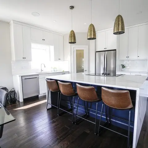 White shaker kitchen with a waterfall-edge marble island, brass dome pendant lights, and stainless built-in double ovens, dining table staged in the foreground.