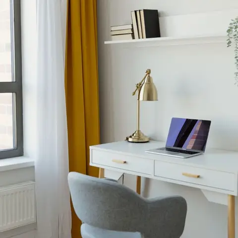 White study desk with a brass desk lamp and laptop, a floating bookshelf with a trailing plant above, and a mustard curtain framing the window