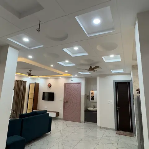 Wide entry passage with a coffered grid false ceiling opening onto a living room with a teal sofa, TV console, and doors to an ensuite bath and bedroom.