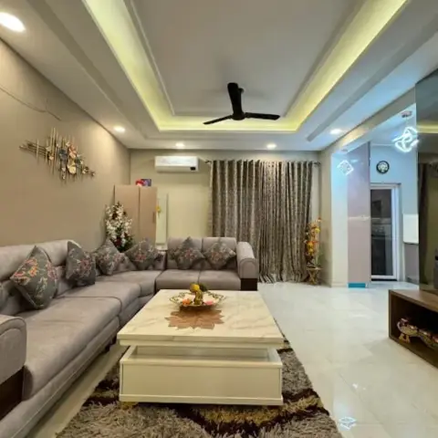 Wide grey L-shaped sofa piled with floral cushions faces a cream coffee table with an inlaid marble top on a brown shag rug