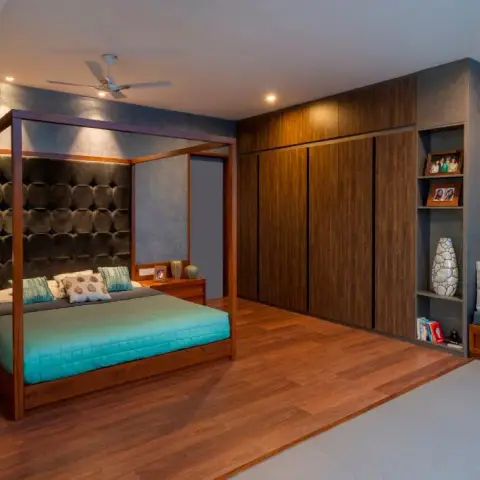 Wide teak four-poster bed against a chocolate tufted-leather headboard panel, flanked by a blue textured wall