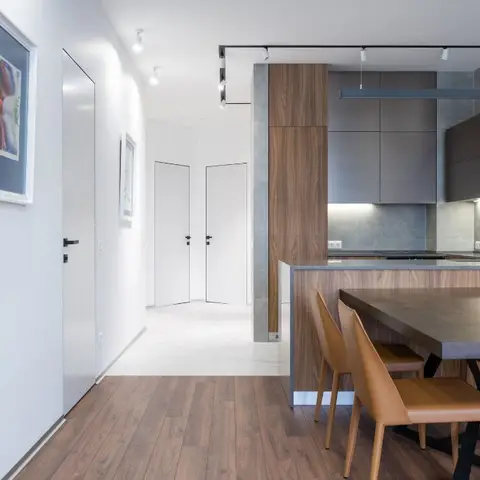 Wood dining table with tan leather chairs beside a matte-grey kitchen island, viewed from a white-doored entrance hallway.