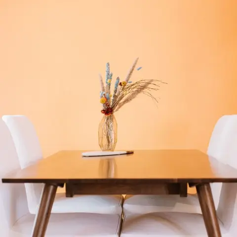 Wood dining table with white upholstered chairs set against a bold orange accent wall, styled with a dried flower arrangement.
