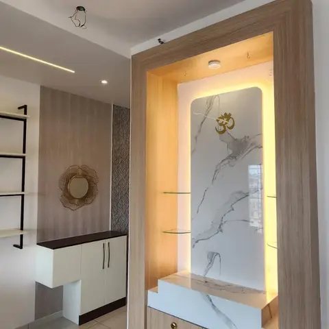 Wood-framed pooja unit with a backlit book-matched marble backdrop, a brass Om motif, glass display shelves, and storage drawers with brass knobs below.