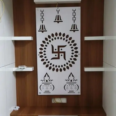 Wood-laminate pooja unit with a white CNC-cut swastik and bell motif panel, floating side shelves and drawer storage below.