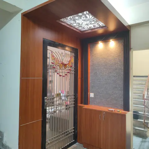Wood-panelled entrance foyer with a backlit floral jaali ceiling cutout above a grille security door and a laminate shoe cabinet under a textured accent wall.