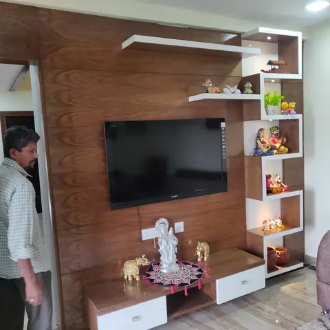 Wood-veneer TV feature wall with a stepped white display shelf showcasing curated brass and clay figurines, beside a jaali partition.