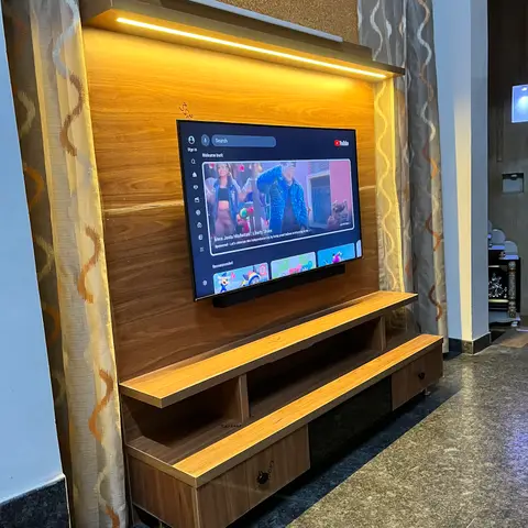 Wood-veneer TV unit with an illuminated floating shelf, open storage cubbies below, and patterned sheer curtains flanking a textured wall.