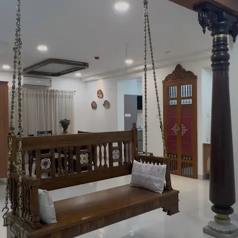 Wooden hanging swing on brass chains in an entrance hall, beside a carved wood pillar and a maroon-and-gold pooja door.
