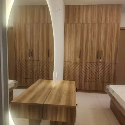 Woodgrain wardrobe run with chevron-grooved lower panels wrapping a corner, a backlit oval mirror cut into a curved white fin and a matching dresser ledge